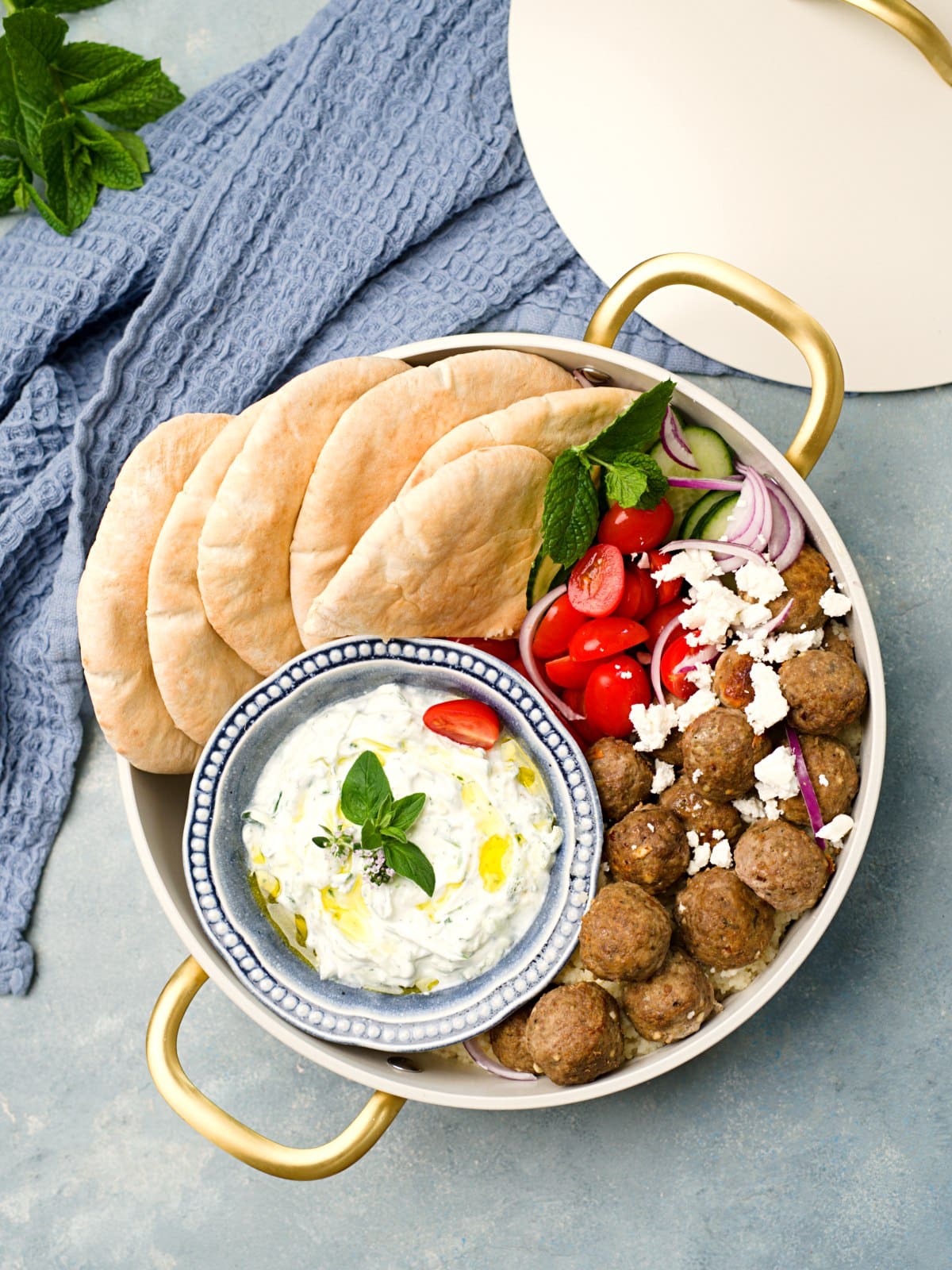 Yläkuva lihapullatarjoilusta valkoisessa kasarissa jonka voi ottaa piknikille, kastiketta, pitaleipiä, kurkkua, tomaattia, sipulia ja kreikkalaisia lihapullia. 